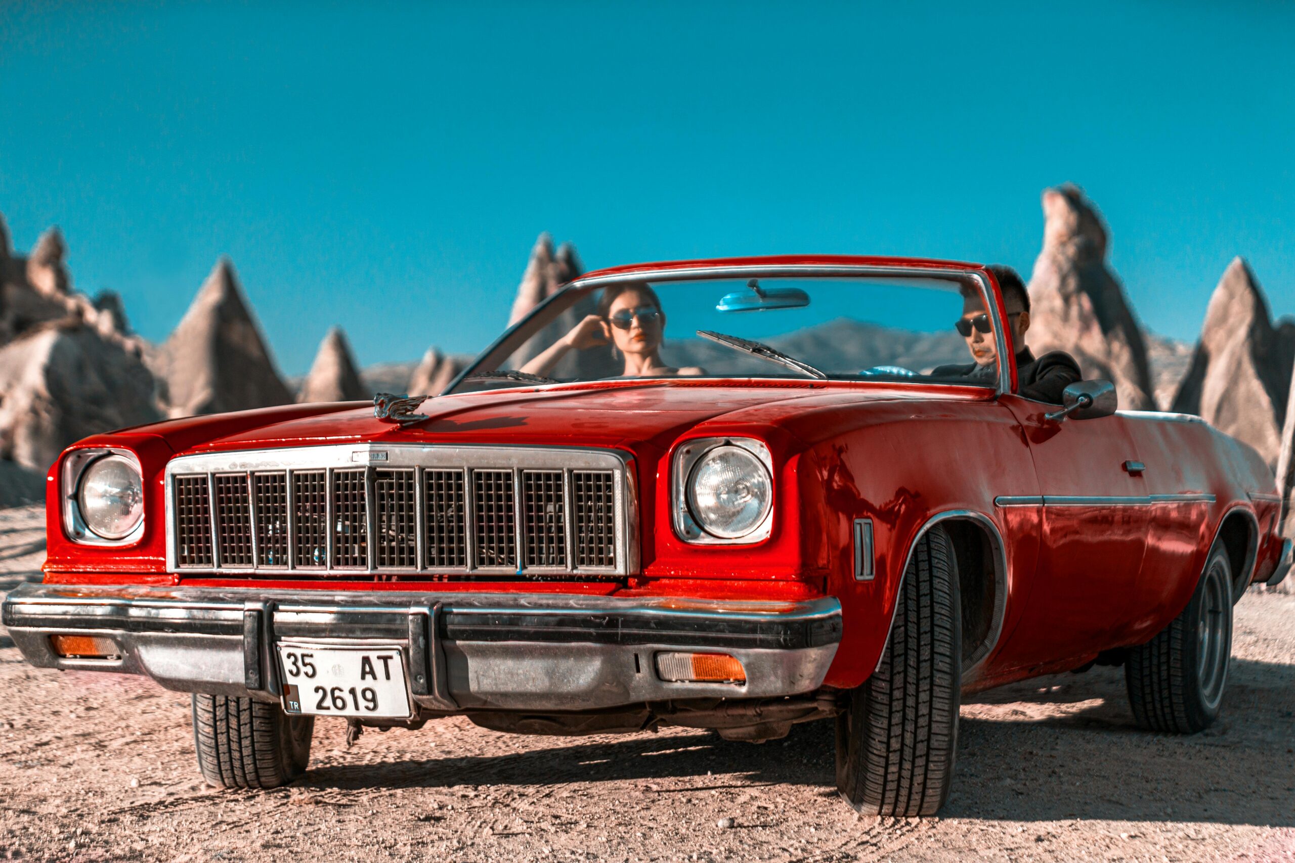 Services Vintage red convertible car with couple in Cappadocia's unique rock formations under a clear blue sky.